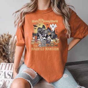 Puede incluir: Camiseta naranja con un gr&aacute;fico de Mickey Mouse y sus amigos frente a la Mansi&oacute;n Encantada en Walt Disney World. El texto "Walt Disney World" y "The Haunted Mansion" est&aacute; impreso en la camiseta.