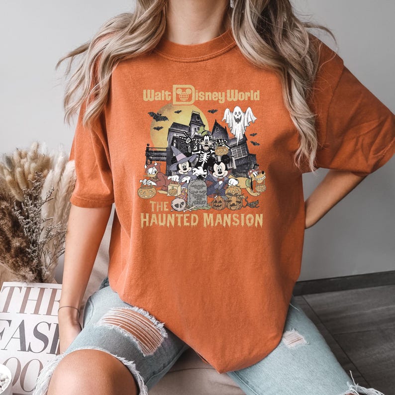 Puede incluir: Camiseta naranja con un gr&aacute;fico de Mickey Mouse y sus amigos frente a la Mansi&oacute;n Encantada en Walt Disney World. El texto "Walt Disney World" y "The Haunted Mansion" est&aacute; impreso en la camiseta.