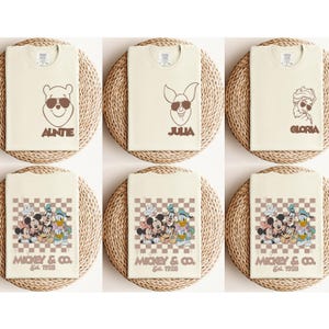 May include: Cream-colored t-shirts with brown graphics of cartoon characters wearing sunglasses. The shirts feature Winnie the Pooh with "AUNTIE", Piglet with "JULIA", and Elsa with "GLORIA". Other shirts display Mickey Mouse, Donald Duck, and Goofy with "MICKEY & CO. Est. 1928".