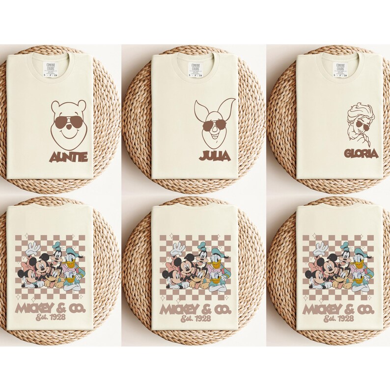 May include: Cream-colored t-shirts with brown graphics of cartoon characters wearing sunglasses. The shirts feature Winnie the Pooh with "AUNTIE", Piglet with "JULIA", and Elsa with "GLORIA". Other shirts display Mickey Mouse, Donald Duck, and Goofy with "MICKEY & CO. Est. 1928".