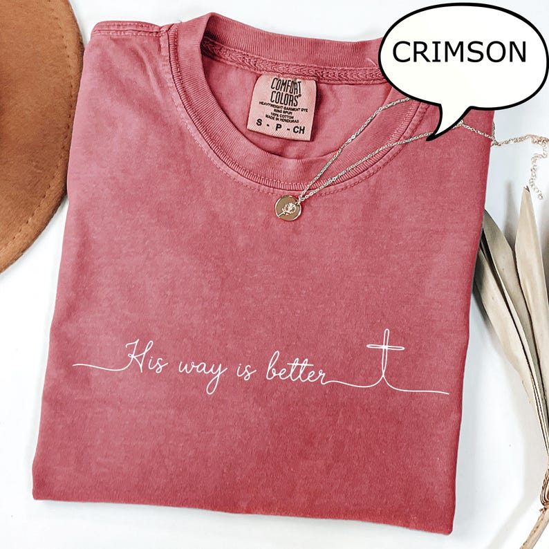 May include: Crimson-colored t-shirt with the text "His way is better" and a cross design. The shirt has a crew neck and a small tag at the top. A gold-colored necklace with a pendant is draped over the shirt.