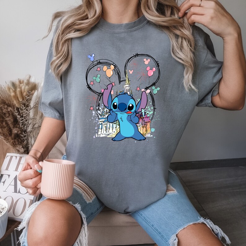 May include: A gray t-shirt with a blue Stitch character holding balloons in front of a cartoon castle. The balloons are pink, blue, green, and yellow. The castle is surrounded by stars.