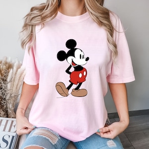 Op de afbeelding: Een roze T-shirt met een cartoon afbeelding van Mickey Mouse in een rood-zwarte outfit. Mickey Mouse glimlacht en heeft zijn hand op zijn heup.