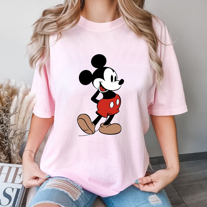 Op de afbeelding: Een roze T-shirt met een cartoon afbeelding van Mickey Mouse in een rood-zwarte outfit. Mickey Mouse glimlacht en heeft zijn hand op zijn heup.