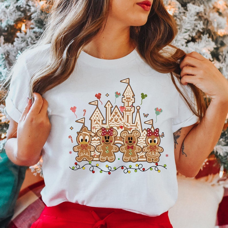 May include: White t-shirt featuring a gingerbread castle and gingerbread characters. The castle is brown with white accents, and the characters are decorated with red bows and candy canes. A string of colorful lights is at the bottom.