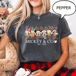 Puede incluir: Camiseta gris oscuro con un dise&ntilde;o navide&ntilde;o con personajes de Disney, como Mickey Mouse y el Pato Donald. La camiseta muestra el texto "Mickey & Co. Est. 1928".