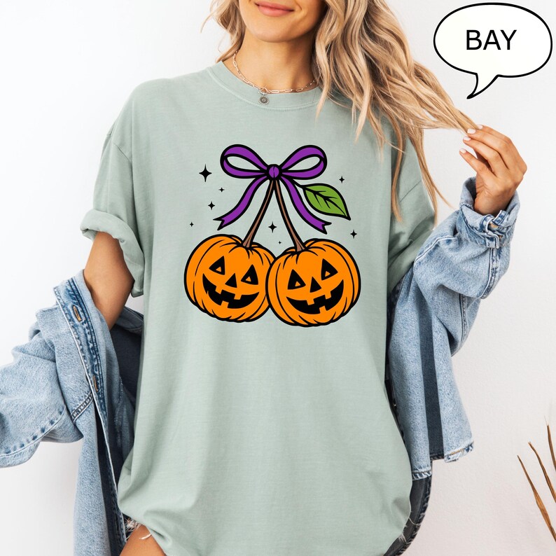 May include: A light green t-shirt featuring a Halloween-themed graphic. The graphic depicts two orange jack-o'-lanterns with black carved faces, connected by a purple bow and green leaf. The word "BAY" is in a speech bubble.