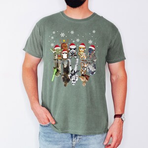 Puede incluir: Una camiseta verde con un dise&ntilde;o de Star Wars con tem&aacute;tica navide&ntilde;a. El dise&ntilde;o presenta un grupo de personajes de Star Wars con gorros de Pap&aacute; Noel y de pie en fila. Los personajes se reflejan en el agua debajo de ellos.