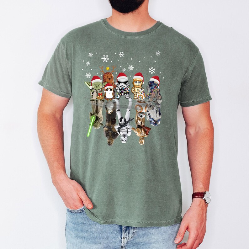 Puede incluir: Una camiseta verde con un dise&ntilde;o de Star Wars con tem&aacute;tica navide&ntilde;a. El dise&ntilde;o presenta un grupo de personajes de Star Wars con gorros de Pap&aacute; Noel y de pie en fila. Los personajes se reflejan en el agua debajo de ellos.