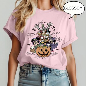 May include: A light pink t-shirt featuring a Halloween-themed graphic. The graphic includes cartoon characters such as Mickey Mouse, Minnie Mouse, Donald Duck, and Goofy, along with a carved pumpkin and bats. The word "BLOSSOM" is in a speech bubble.
