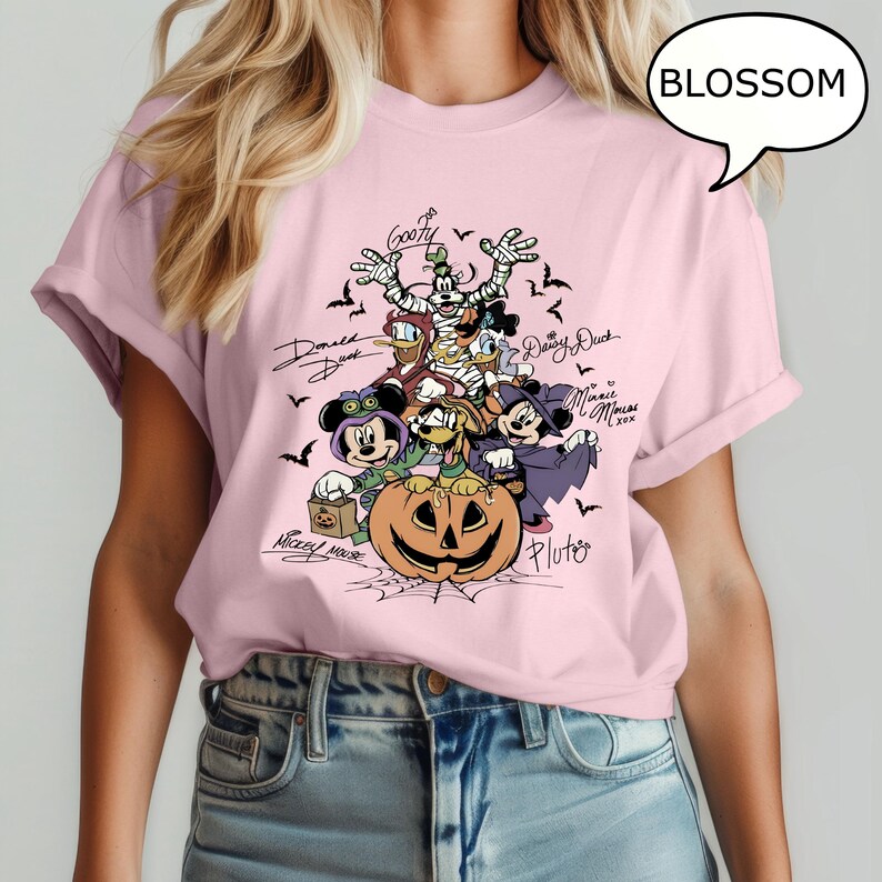 May include: A light pink t-shirt featuring a Halloween-themed graphic. The graphic includes cartoon characters such as Mickey Mouse, Minnie Mouse, Donald Duck, and Goofy, along with a carved pumpkin and bats. The word "BLOSSOM" is in a speech bubble.