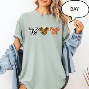 Pu&ograve; includere: T-shirt verde chiaro con tre forme di testa di Topolino con motivi animalier: zebra, leopardo e giraffa. La parola "BAY" &egrave; in una nuvoletta. La maglietta ha le maniche arrotolate.