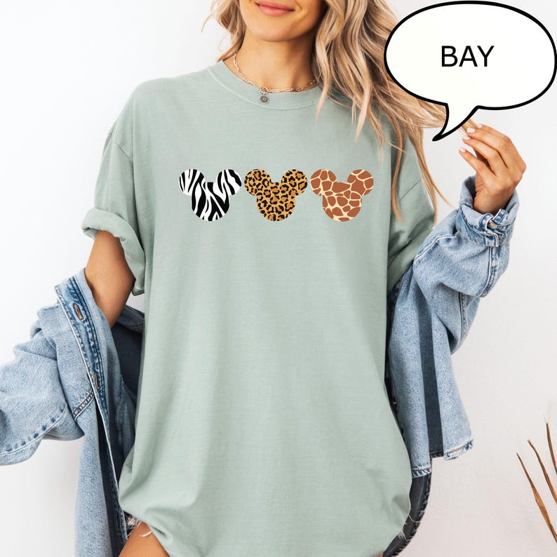 Pu&ograve; includere: T-shirt verde chiaro con tre forme di testa di Topolino con motivi animalier: zebra, leopardo e giraffa. La parola "BAY" &egrave; in una nuvoletta. La maglietta ha le maniche arrotolate.