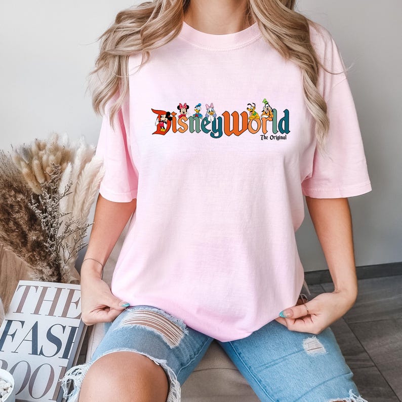 K&ouml;nnte beinhalten: Ein pinkfarbenes T-Shirt mit einem Aufdruck von Disney World-Charakteren und dem Text "Disney World The Original".