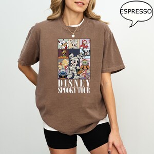 May include: Espresso-colored t-shirt featuring a grid of Disney characters in Halloween costumes. The shirt has the text "DISNEY SPOOKY TOUR" printed below the grid. The shirt is a relaxed fit.