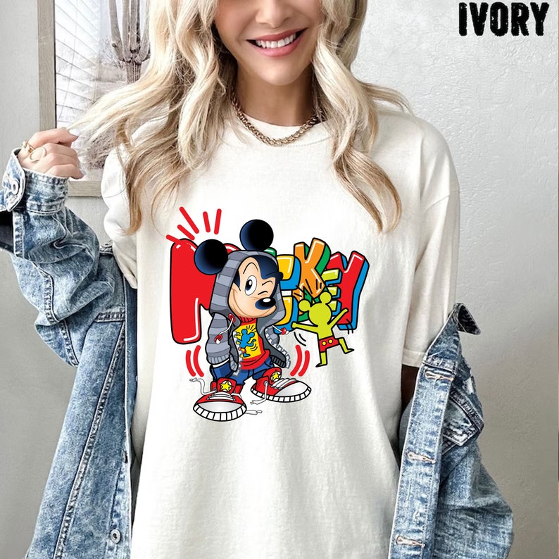 Pode incluir: Camiseta branca com um gr&aacute;fico colorido do Mickey Mouse vestindo um moletom com capuz e t&ecirc;nis vermelhos. O gr&aacute;fico tamb&eacute;m inclui a palavra "Mickey" em letras coloridas.