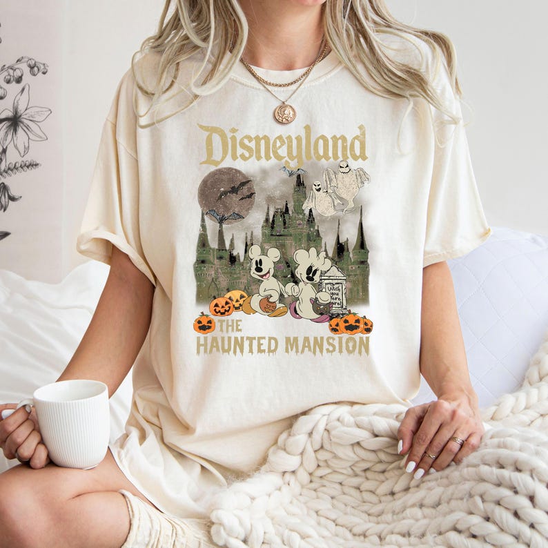 May include: A cream-colored t-shirt with a vintage-style graphic of Mickey Mouse and a ghost in front of the Haunted Mansion at Disneyland. The text "Disneyland" and "The Haunted Mansion" are printed on the shirt. Pumpkins are scattered around the bottom of the graphic.