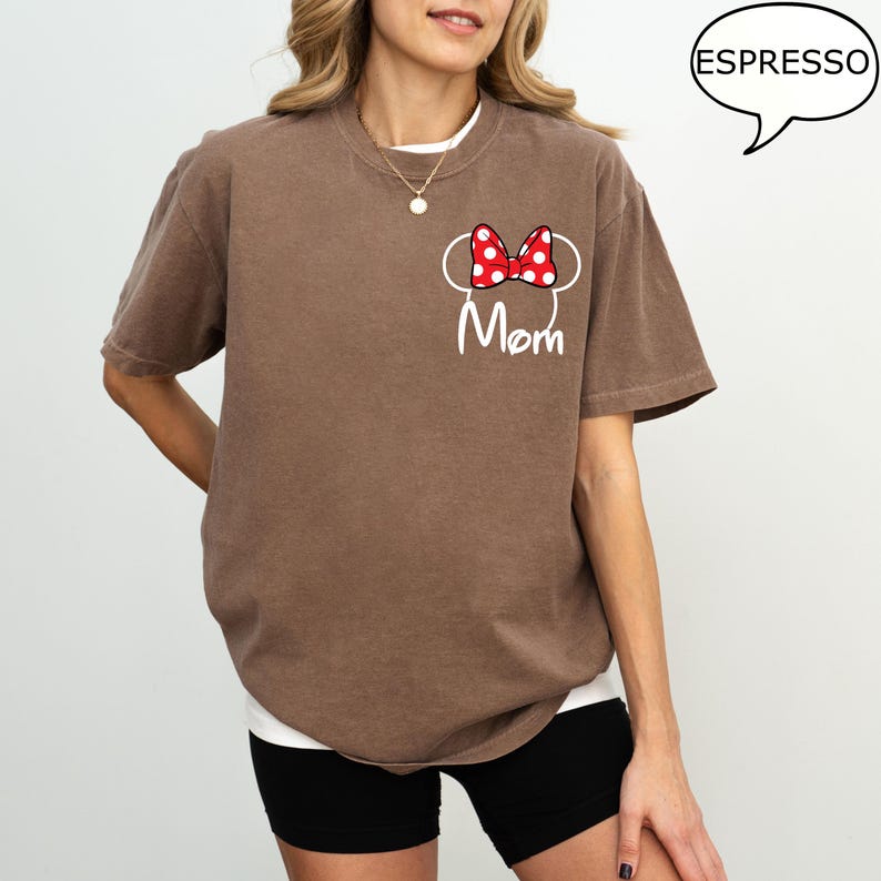 May include: Brown t-shirt with a white graphic of Minnie Mouse ears and a red polka dot bow. The word "Mom" is written in white script below the graphic. The shirt is a casual style.