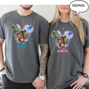 May include: Two gray t-shirts with a colorful Disney-themed design. The design features characters from the movie Moana, surrounded by flowers, in a Mickey Mouse head shape. One shirt says "DAD" in blue, the other says "MOM" in pink.