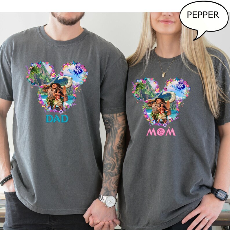 May include: Two gray t-shirts with a colorful Disney-themed design. The design features characters from the movie Moana, surrounded by flowers, in a Mickey Mouse head shape. One shirt says "DAD" in blue, the other says "MOM" in pink.