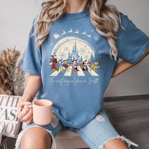 Puede incluir: Una camiseta azul con un dise&ntilde;o navide&ntilde;o que presenta a Mickey Mouse y sus amigos caminando por un paso de cebra frente al Castillo de Cenicienta. El texto "The most magical place on Earth" est&aacute; impreso debajo del dise&ntilde;o.