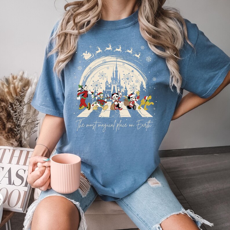 Puede incluir: Una camiseta azul con un dise&ntilde;o navide&ntilde;o que presenta a Mickey Mouse y sus amigos caminando por un paso de cebra frente al Castillo de Cenicienta. El texto "The most magical place on Earth" est&aacute; impreso debajo del dise&ntilde;o.