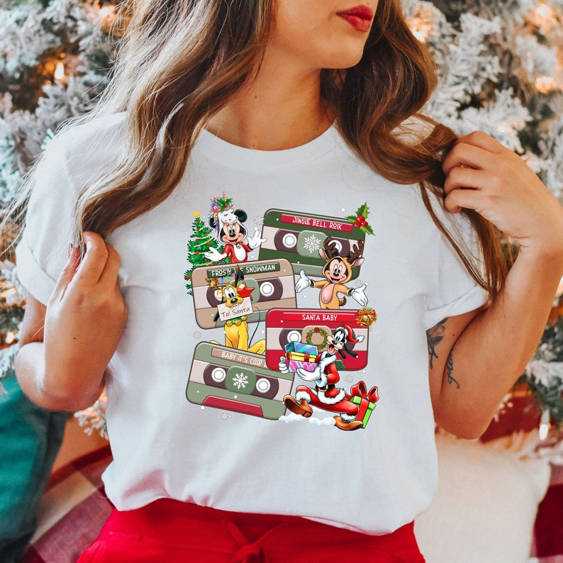 Puede incluir: Camiseta blanca con un dise&ntilde;o navide&ntilde;o con personajes de dibujos animados de Disney. El dise&ntilde;o incluye cinco cintas de cassette con diferentes canciones navide&ntilde;as impresas en ellas. Los personajes incluyen Mickey Mouse, Pluto, Goofy y Donald Duck.