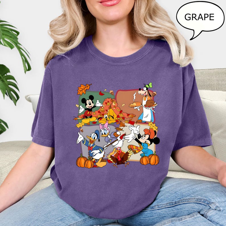 May include: A purple t-shirt featuring a colorful graphic of Disney characters celebrating autumn. Mickey Mouse, Donald Duck, and Goofy are among the characters depicted with pumpkins and fall leaves. The word "GRAPE" is in a speech bubble.