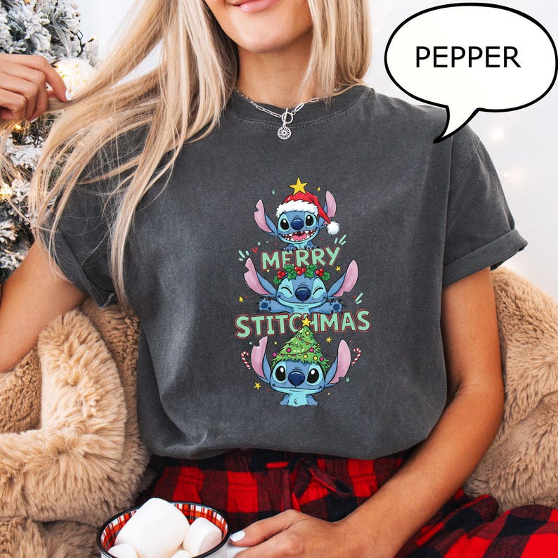 May include: Dark gray t-shirt featuring a stacked design of the character Stitch from Lilo & Stitch, with a Santa hat, Christmas tree, and the words "MERRY STITCHMAS" in green and white. The shirt is a comfortable, casual style.