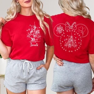 K&ouml;nnte beinhalten: Zwei rote T-Shirts mit wei&szlig;en Designs. Ein Shirt zeigt eine Mickey-Maus-Silhouette mit dem Text "The Happiest Place on Earth" und Feuerwerk. Das andere Shirt zeigt eine Mickey-Maus-Silhouette mit dem Text "Jessie" und ein Schloss.