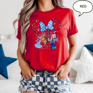 Puede incluir: Una camiseta roja con un dise&ntilde;o inspirado en Minnie Mouse, que incluye un lazo azul y fuegos artificiales. El dise&ntilde;o incorpora un castillo y varias princesas de Disney. La palabra "RED" est&aacute; en un bocadillo.