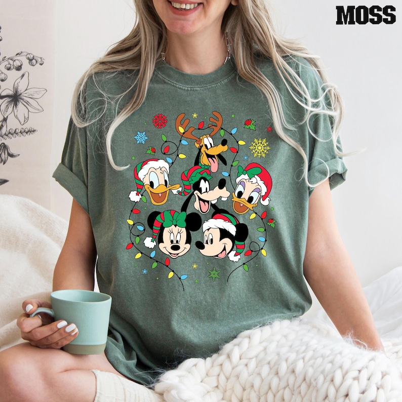May include: A green t-shirt with a Christmas-themed design featuring Mickey Mouse, Minnie Mouse, Donald Duck, Goofy, and Pluto. The characters are surrounded by colorful Christmas lights.