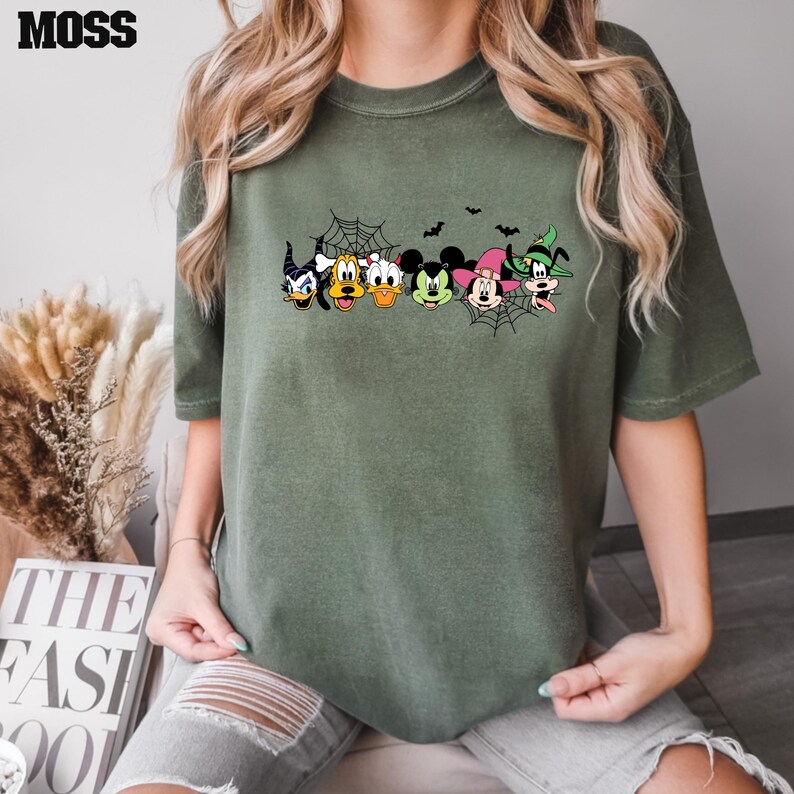 May include: A green t-shirt with a Halloween-themed design featuring Mickey Mouse, Minnie Mouse, Donald Duck, Goofy, and Pluto. The characters are dressed in costumes and are surrounded by bats and a spider web.