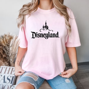 Puede incluir: Camiseta rosa claro con la palabra "Disneyland" en una fuente estilizada y un castillo negro encima. La camiseta de manga corta es de color liso.