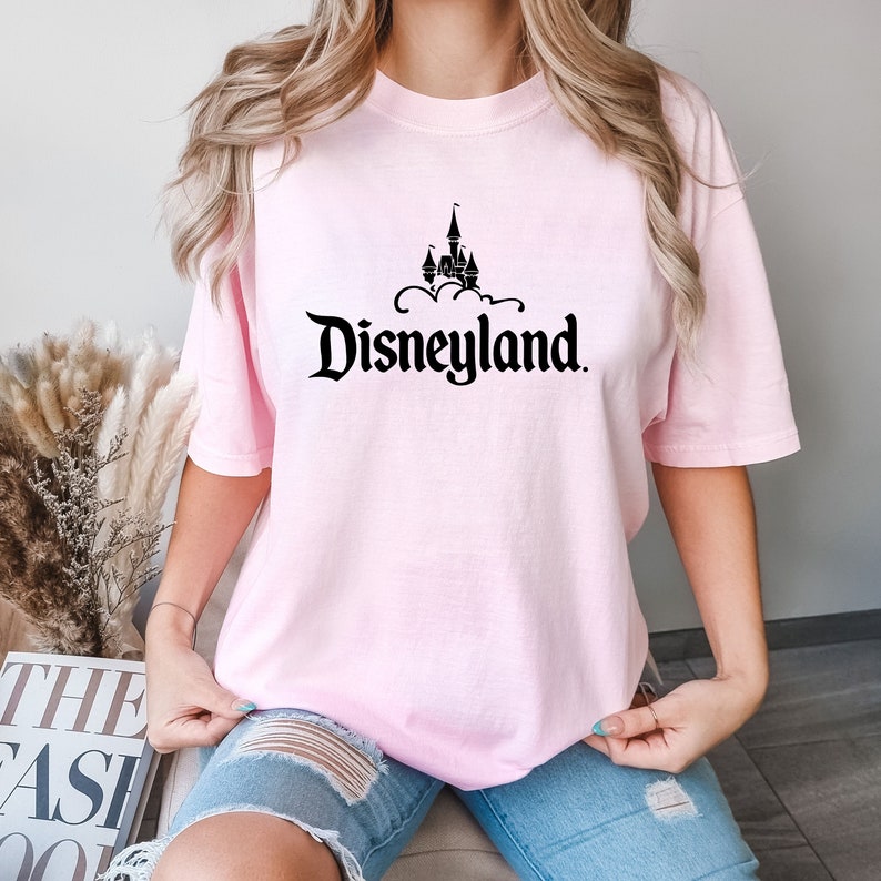 Puede incluir: Camiseta rosa claro con la palabra "Disneyland" en una fuente estilizada y un castillo negro encima. La camiseta de manga corta es de color liso.