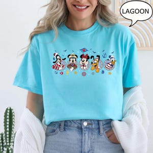 May include: A light blue t-shirt featuring a nautical-themed graphic with cartoon characters. The graphic includes Goofy, Mickey Mouse, Minnie Mouse, Pluto, and Donald Duck, each in sailor attire. The word "LAGOON" is in a speech bubble.