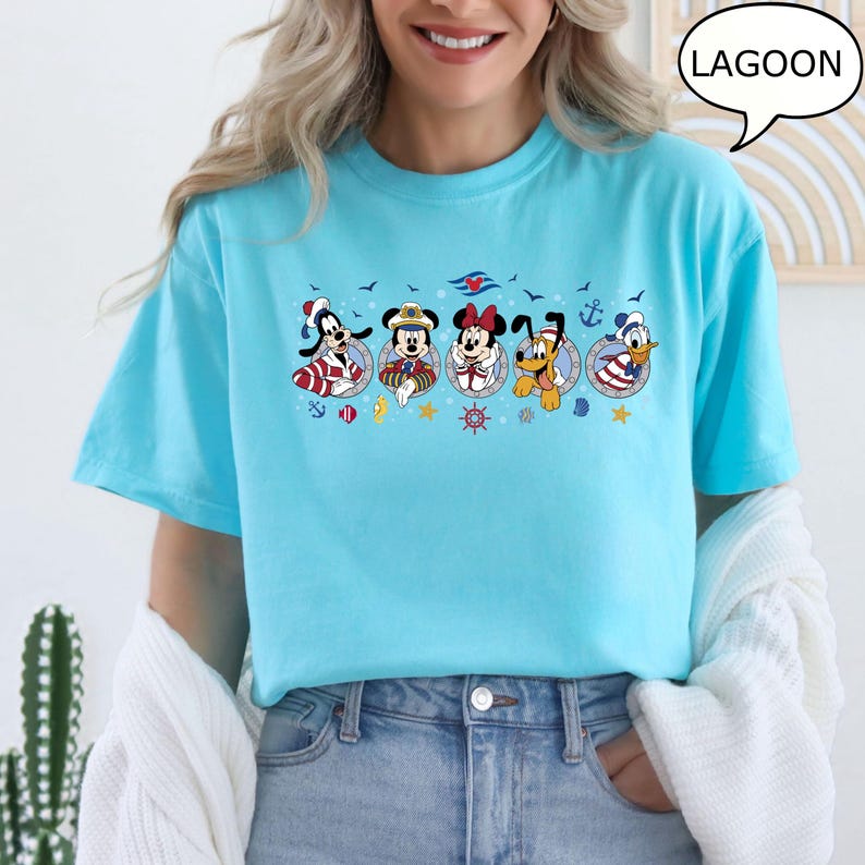 May include: A light blue t-shirt featuring a nautical-themed graphic with cartoon characters. The graphic includes Goofy, Mickey Mouse, Minnie Mouse, Pluto, and Donald Duck, each in sailor attire. The word "LAGOON" is in a speech bubble.