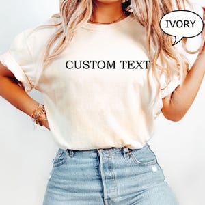 May include: A white t-shirt with the text "CUSTOM TEXT" printed on the front. The t-shirt is being worn by a person and is available in the color ivory.