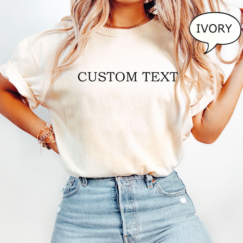 May include: A white t-shirt with the text "CUSTOM TEXT" printed on the front. The t-shirt is being worn by a person and is available in the color ivory.