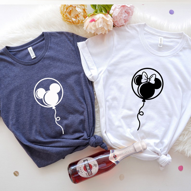 Pode incluir: Duas camisetas com um design de bal&atilde;o do Mickey Mouse e da Minnie Mouse. O bal&atilde;o do Mickey Mouse &eacute; branco em uma camiseta azul escura. O bal&atilde;o da Minnie Mouse &eacute; preto em uma camiseta branca.