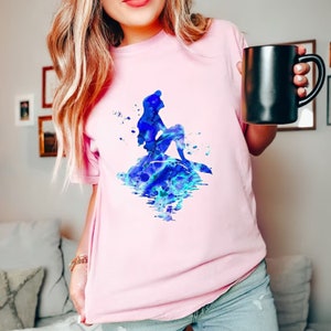 Puede incluir: Una camiseta rosa con una ilustraci&oacute;n de acuarela azul de Ariel, la Sirenita, sentada en una roca con su cola en el agua.