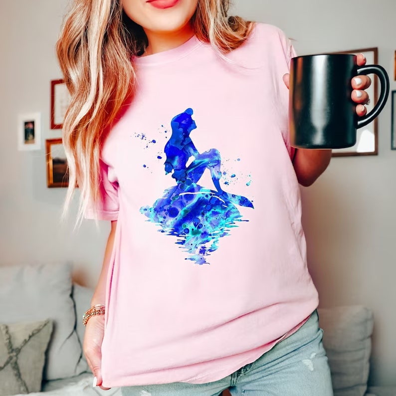 Puede incluir: Una camiseta rosa con una ilustraci&oacute;n de acuarela azul de Ariel, la Sirenita, sentada en una roca con su cola en el agua.