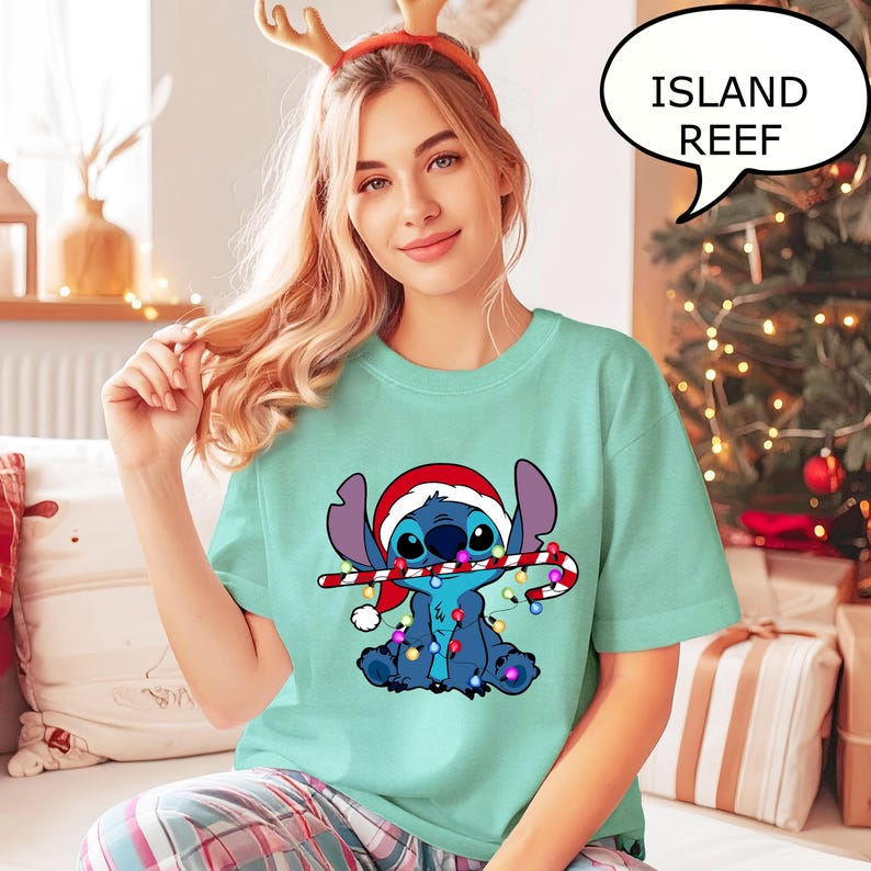 Puede incluir: Una camiseta verde azulado claro con un personaje de dibujos animados, Stitch, que lleva un gorro de Pap&aacute; Noel y sostiene un bast&oacute;n de caramelo. El personaje est&aacute; rodeado de luces navide&ntilde;as. El texto "ISLAND REEF" est&aacute; en una burbuja.