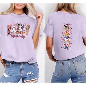 May include: A light purple t-shirt featuring the text "HAPPY Valentine's Day" with Disney characters. The back of the shirt has a vertical design with more Disney characters and hearts. The shirt is paired with blue jeans.