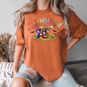 Puede incluir: Camiseta naranja con un gr&aacute;fico de Mickey Mouse, Minnie Mouse, Goofy, Donald Duck y Pluto vestidos con disfraces de Halloween. El texto "Disneyland" est&aacute; por encima de los personajes.
