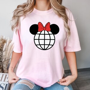 K&ouml;nnte beinhalten: Ein pinkfarbenes T-Shirt mit einem schwarzen und wei&szlig;en Globus-Motiv, auf dem ein rotes Band sitzt, das an Mickey Mouse-Ohren erinnert.