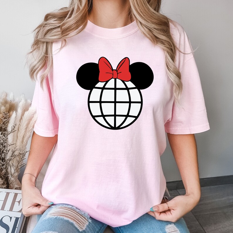 K&ouml;nnte beinhalten: Ein pinkfarbenes T-Shirt mit einem schwarzen und wei&szlig;en Globus-Motiv, auf dem ein rotes Band sitzt, das an Mickey Mouse-Ohren erinnert.