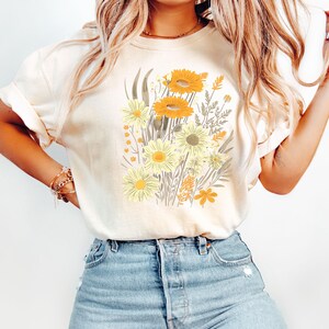 May include: Cream-colored t-shirt featuring a floral design with yellow and orange flowers, green leaves, and brown stems. The graphic is centered on the front of the shirt. The shirt has short sleeves and a relaxed fit, suitable for casual wear.