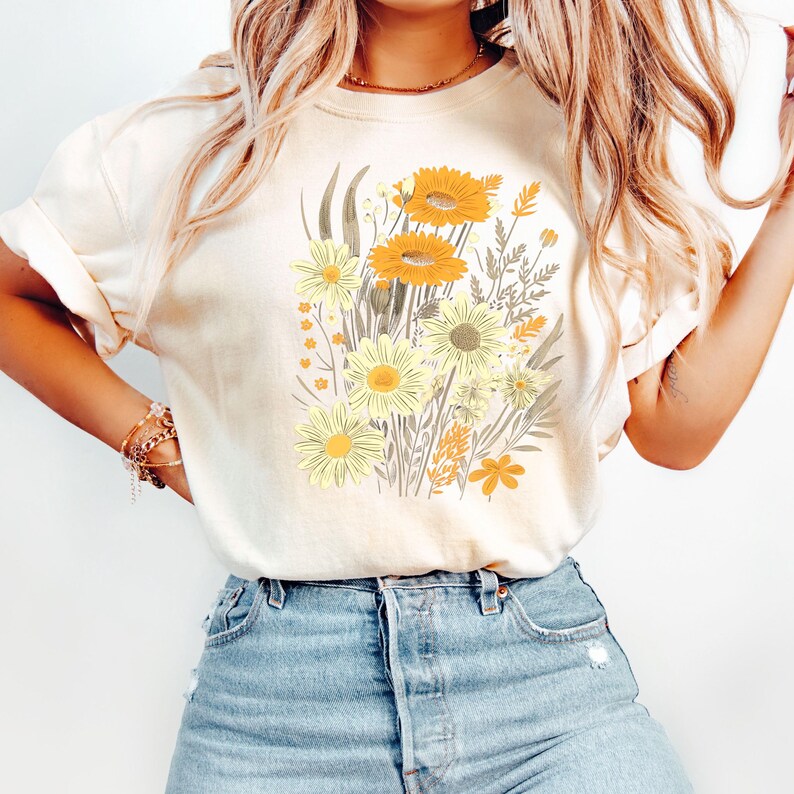 May include: Cream-colored t-shirt featuring a floral design with yellow and orange flowers, green leaves, and brown stems. The graphic is centered on the front of the shirt. The shirt has short sleeves and a relaxed fit, suitable for casual wear.