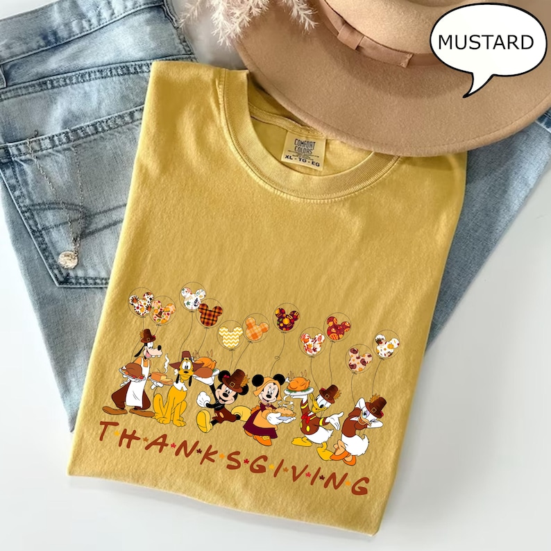 May include: Mustard-colored t-shirt with a Thanksgiving-themed graphic featuring cartoon characters and balloons. The text "THANKSGIVING" is printed across the bottom. A tan hat and blue jeans are also in the image.
